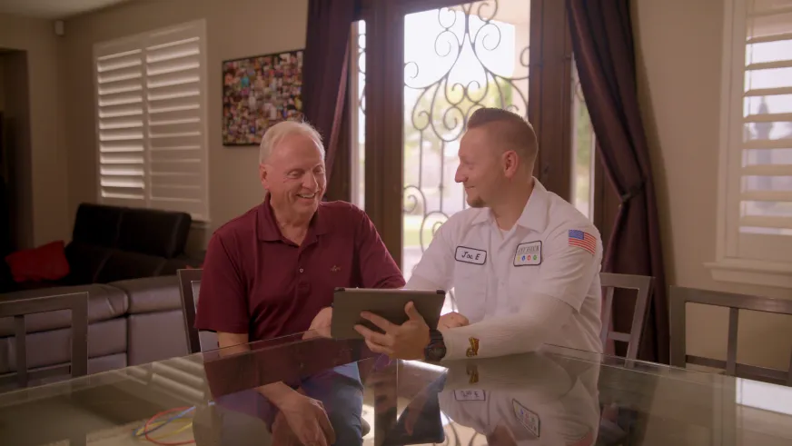 A technician in a white uniform shows an elderly man a document on a tablet in a bright kitchen. Both are engaged and smiling, conveying friendliness.