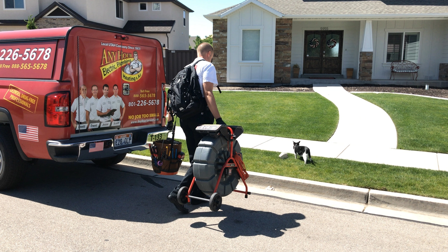 A plumbing technician walks toward a house, pulling equipment from a branded service truck. A black and white cat sits on the lawn nearby.