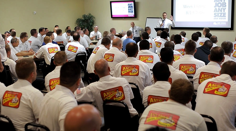 A large group of employees in uniformed shirts with logos attend a meeting in a conference room. A speaker presents at the front, creating a focused atmosphere.