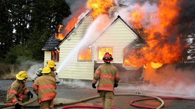 Firefighters in gear spray water on a house engulfed in bright orange flames and heavy smoke, conveying urgency and intense action.