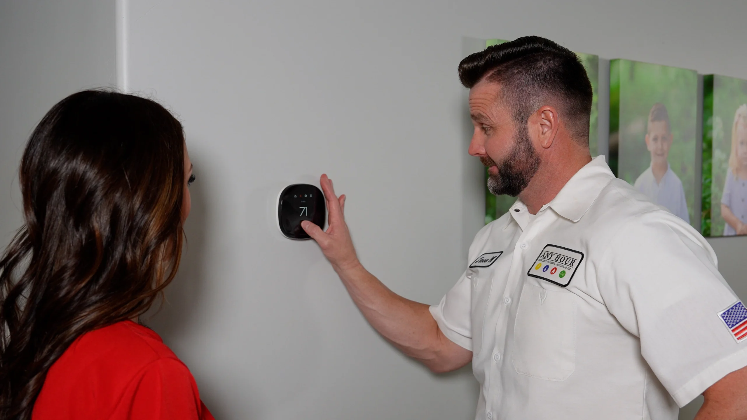 A technician in uniform demonstrates a smart thermostat to a woman in a red shirt. They stand against a light gray wall with children's photos, discussing settings.