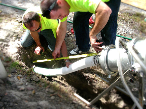 Two men in bright shirts measure a white pipe using a measuring tape. They are kneeling in a dug-out section of earth, focused and working collaboratively.