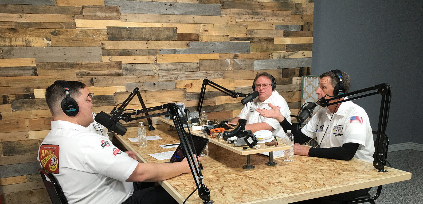 Three men in white shirts engage in a podcast discussion. They're seated around a wooden table with microphones, headsets, and audio equipment. The setting is casual and focused.