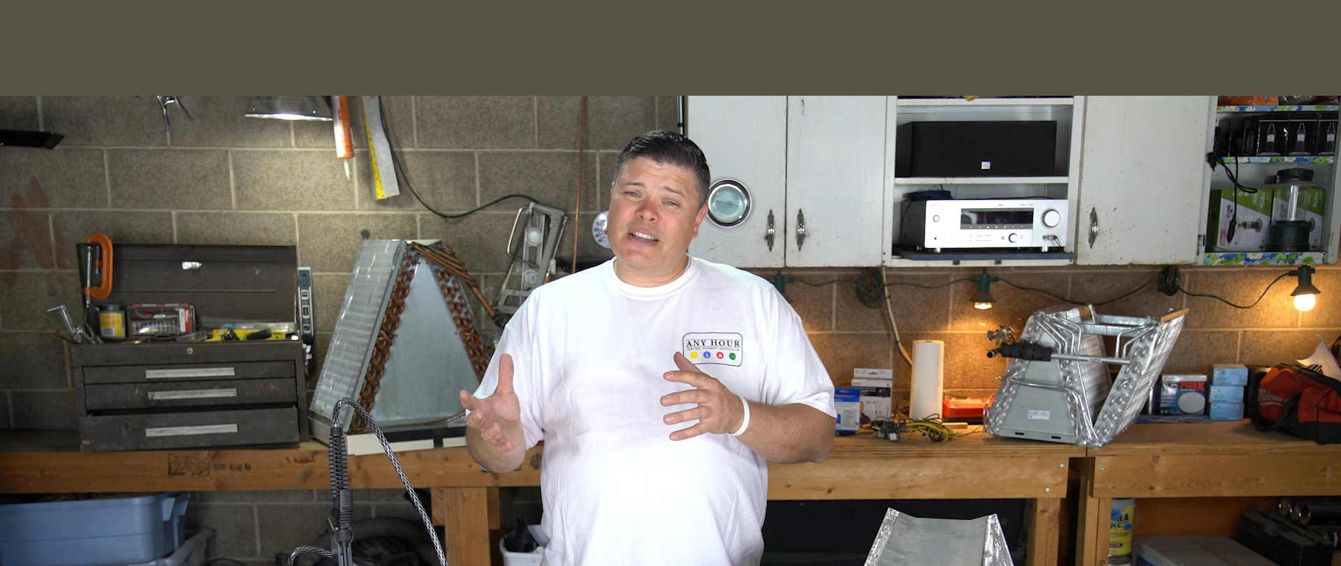 Man in a workshop wearing a white t-shirt stands in front of a workbench with tools, machinery, and cabinets. Bright, organized environment.