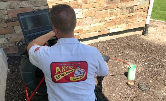 A technician in a branded shirt inspects a laptop connected to plumbing equipment outdoors. Brick wall and mulch are in the background. Focused, professional tone.