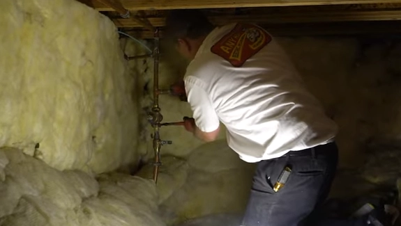 A person in a white shirt works on plumbing in a dimly lit crawl space. The surrounding walls are lined with yellow insulation. The scene is focused and industrious.