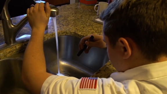 A person examines a running kitchen faucet with a flashlight, their arm resting on a counter. They wear a shirt with a U.S. flag patch. The setting is a cozy kitchen.