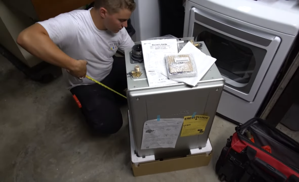 A technician measures a new water heater in a laundry room, surrounded by a dryer and toolbox. The scene conveys careful preparation and focus.