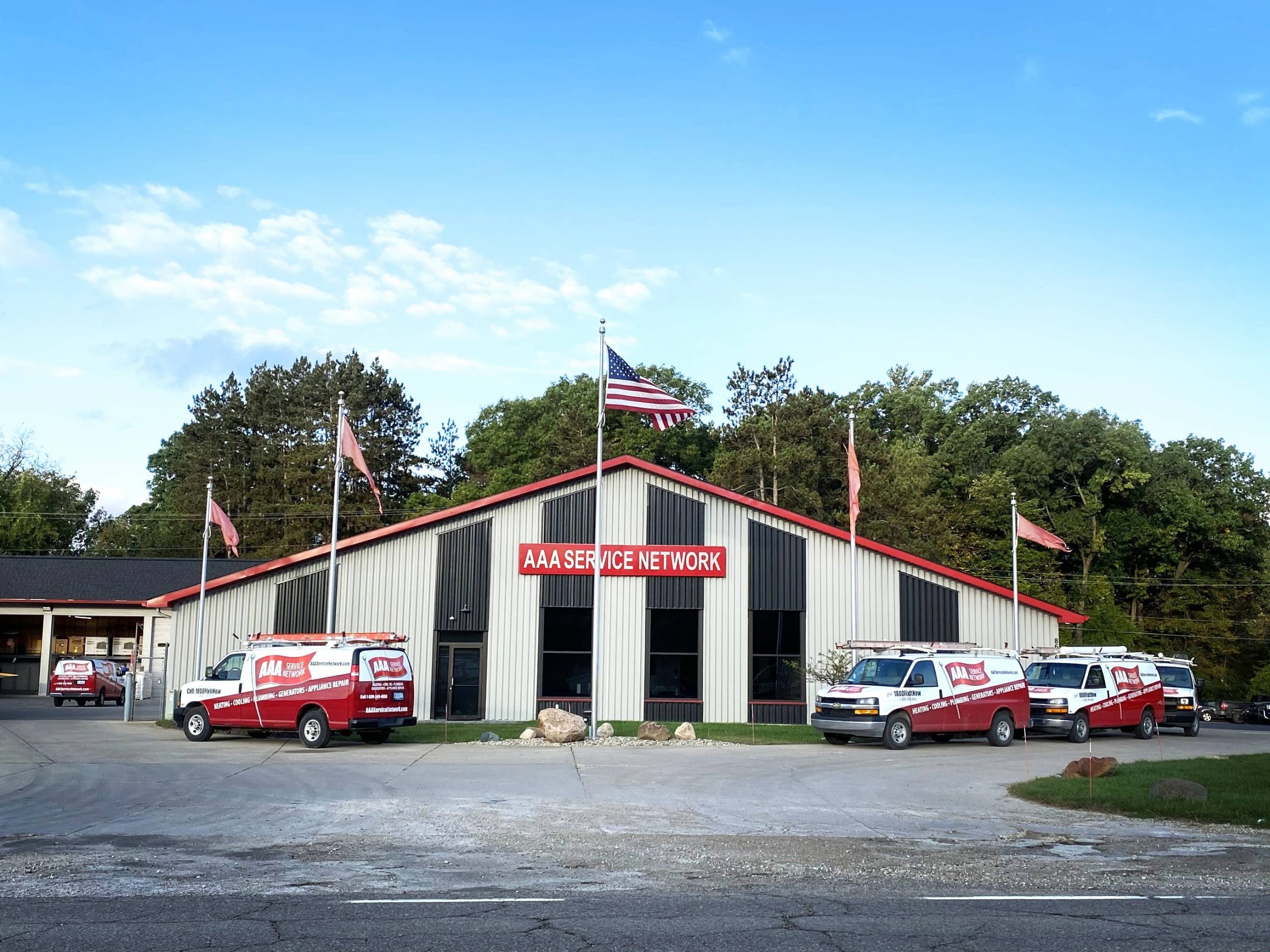 A white and red building with a sign reading AAA Service Network. Several branded vans are parked in front. The scene is calm, with a clear, blue sky and trees in the background. An American flag is raised above the building.