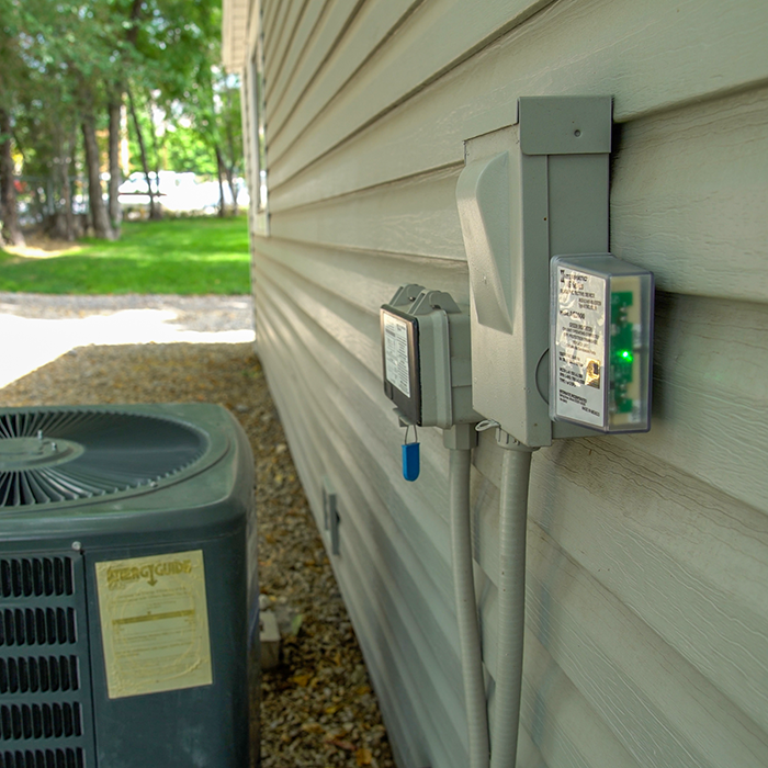 Outdoor electrical boxes are mounted on a light gray house siding. A surge protective device, the AG3000, has a glowing green indicator light.