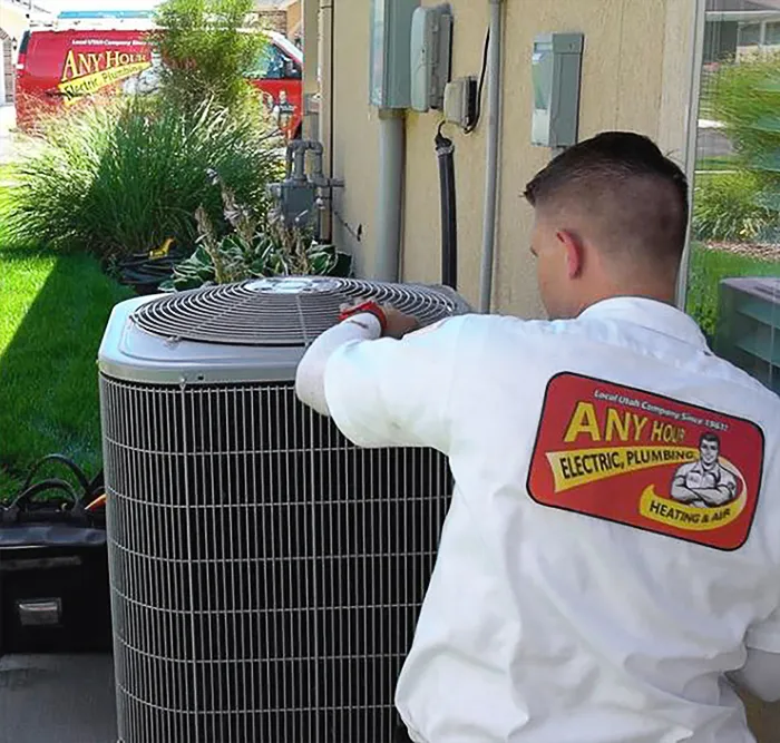 A technician in a white uniform works on an outdoor air conditioning unit. The shirt displays a company logo. A service van is visible in the background.