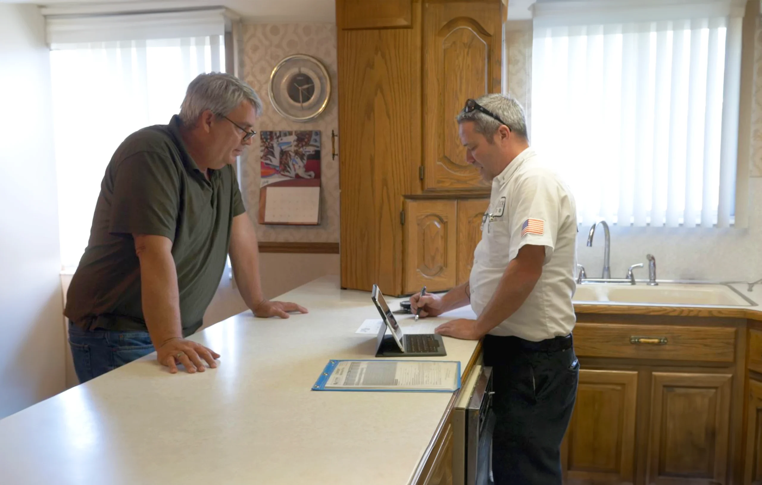 A customer and repair technician discuss repair details in a kitchen. The technician is writing on a tablet, while the customer leans on the counter, looking engaged.