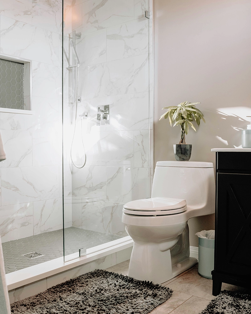 Modern bathroom with a white toilet beside a sleek black vanity. A small potted plant sits on the toilet. The glass shower has marble tile walls.