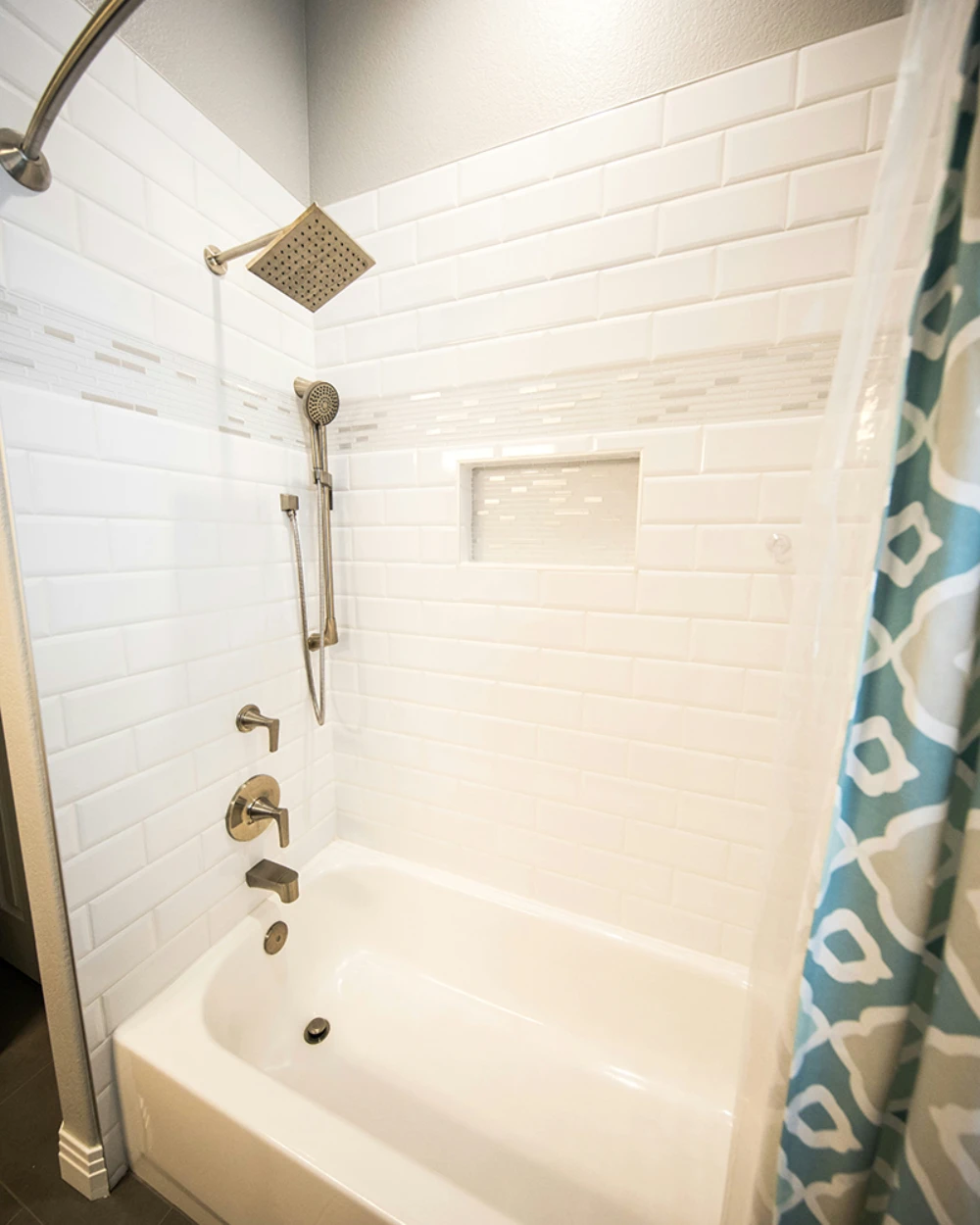 A modern bathroom features a white bathtub with shiny silver fixtures, a square rain showerhead, white subway tile walls, and a teal patterned shower curtain.