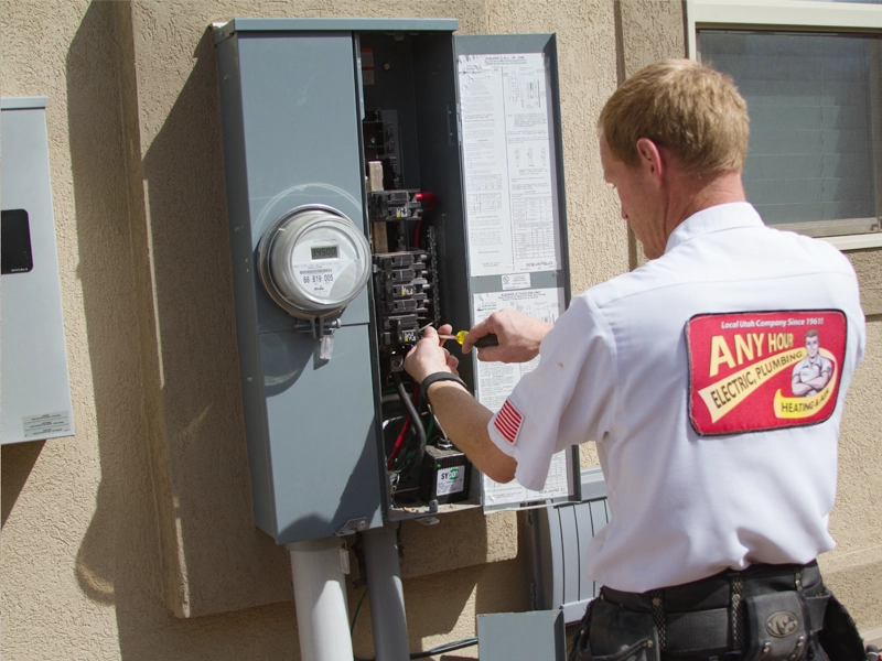 Electrician working on an open gray electrical panel outside a building. He's wearing a uniform with company branding, focused on wiring tasks.