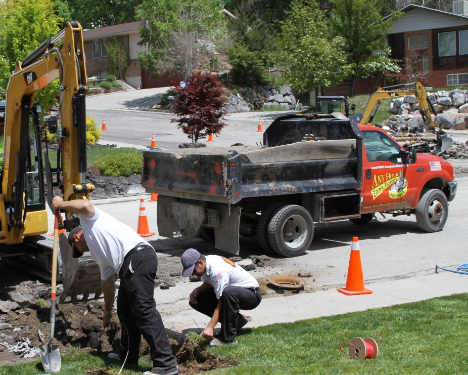 Two workers in a residential area dig near a small excavator and dump truck. Orange cones line the street, indicating roadwork in progress.