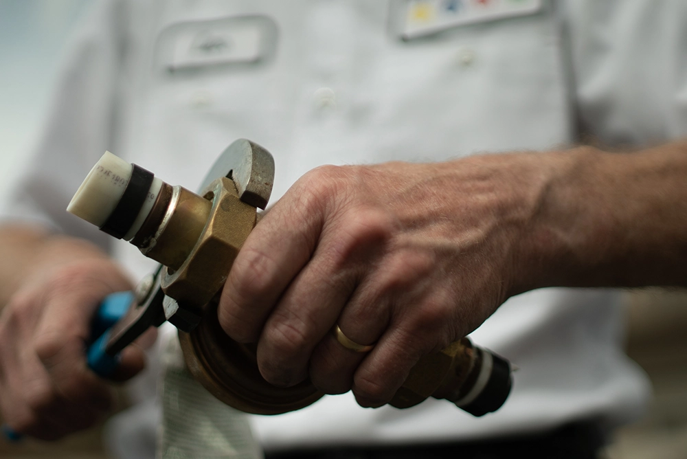 Close-up of a person's hands holding a brass plumbing valve, using a tool to fix it. They wear a white uniform, suggesting a professional setting.