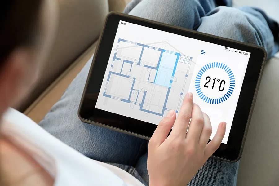 A person holds a tablet displaying a digital home floor plan and a large thermostat reading of 21°C. The scene conveys modern smart home technology.