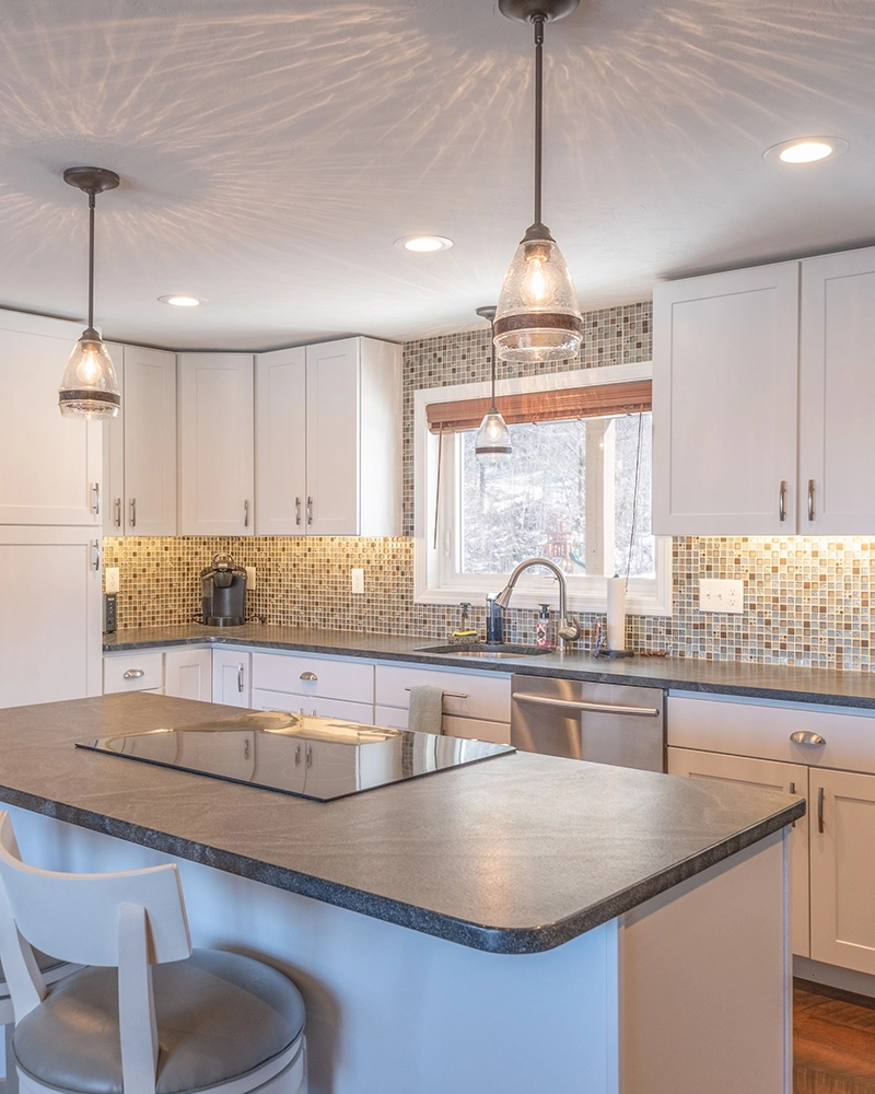 Modern kitchen with white cabinets, mosaic tile backsplash, and dark countertops. Pendant lights hang above a central island with a stovetop, creating a warm ambiance.