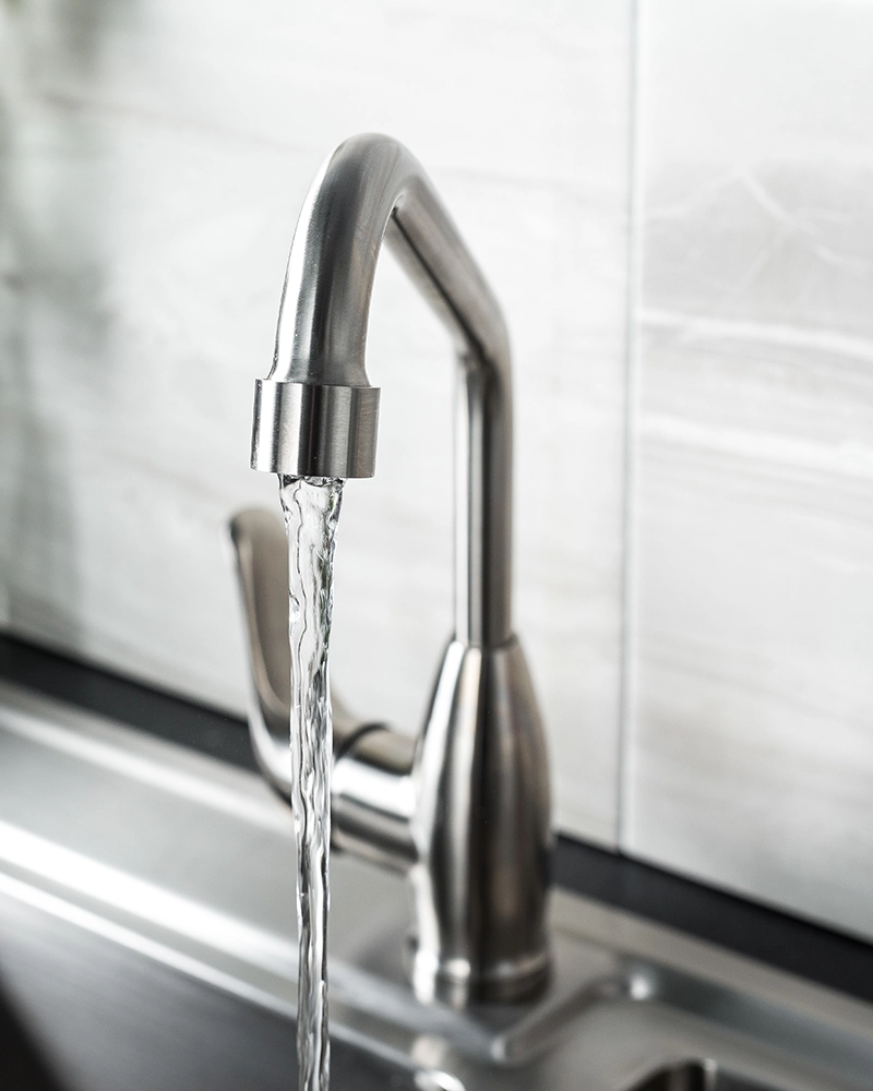 A sleek stainless steel kitchen faucet with a modern design, releasing a steady stream of water into a silver sink against a white tiled backsplash.