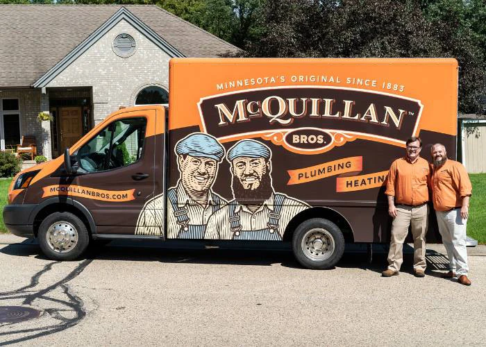 Two men in matching orange shirts stand beside a McQuillan Bros. plumbing van. The van's design includes illustrations of two workers. The mood is professional and cheerful.