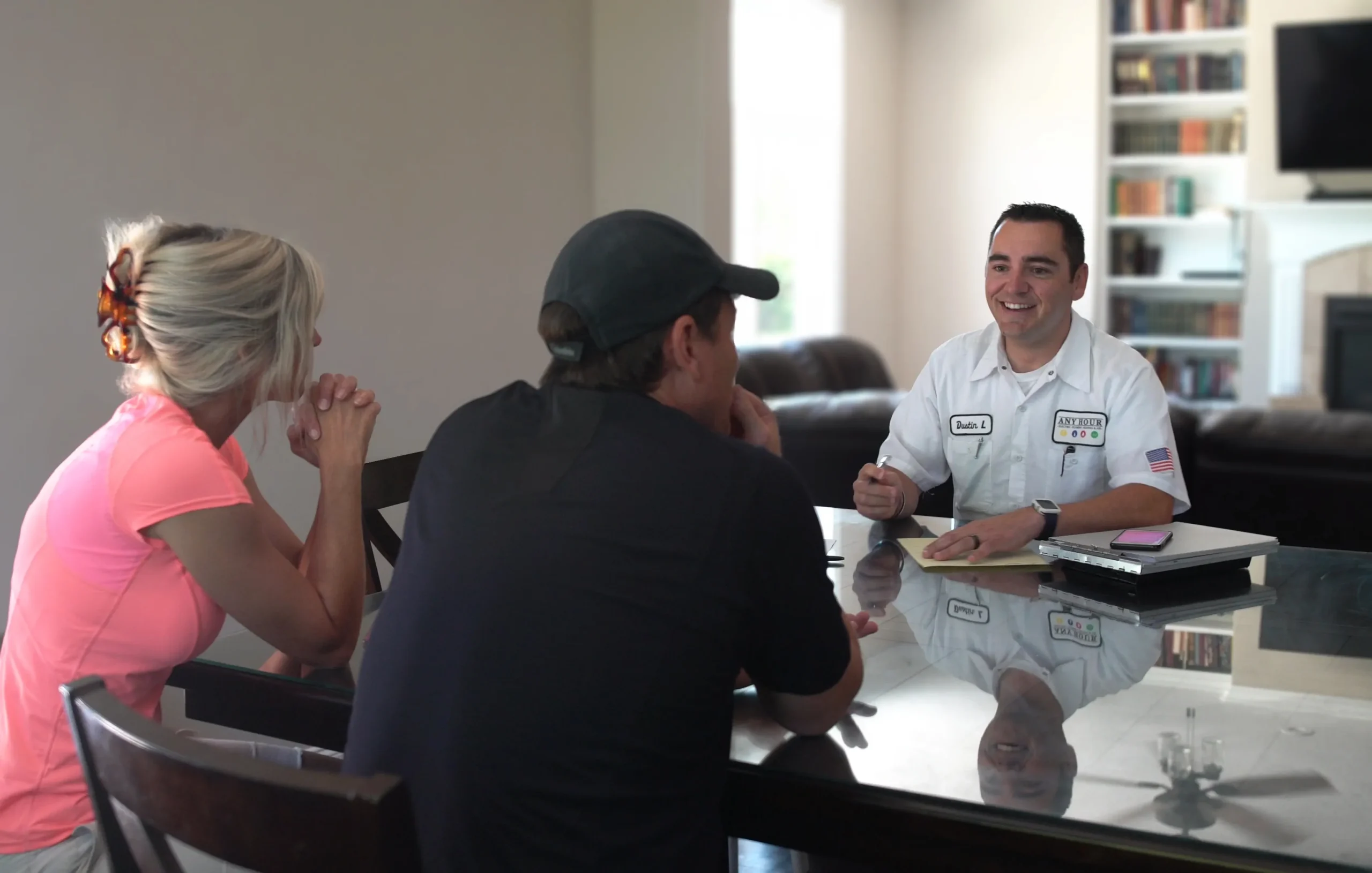 A technician in a white uniform sits at a table, smiling and holding a pen, talking to a couple. The setting is a cozy living room with bookshelves.