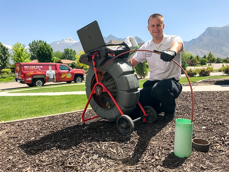 A worker in a white uniform kneels beside sewer inspection equipment on a landscaped lawn. A red service van is in the background, with mountains under a clear blue sky.