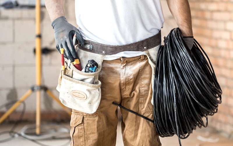 A construction worker in a white shirt and gloves holds a large coil of black cable. He wears a tool belt with various tools, standing in a brick-walled room.