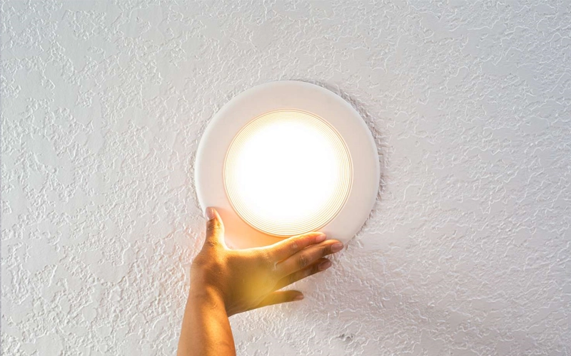 A hand installing a bright, circular ceiling light on a textured white ceiling. The scene conveys warmth and household lighting.