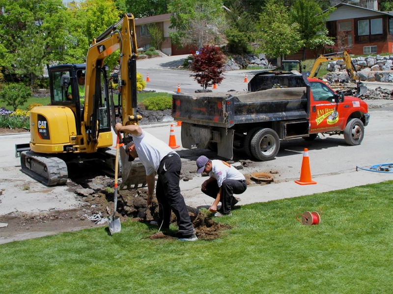 Two workers repair a sidewalk using shovels, near a small excavator and a dump truck. Orange cones mark the area, with residential homes in the background.