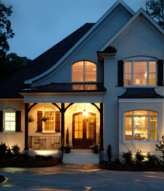 Elegant house at dusk with warm lighting illuminating the arched entryway. Large windows glow, highlighting the inviting and cozy atmosphere.