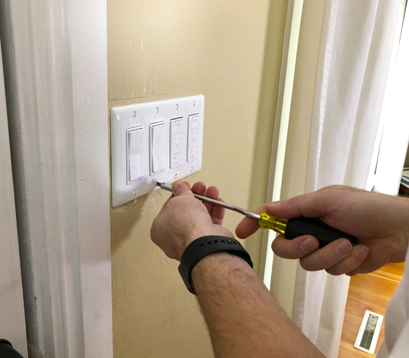 A electrician uses a screwdriver to adjust a white light switch on a beige wall. The scene suggests home maintenance.