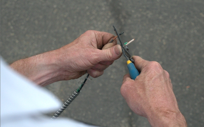 Close-up of hands using wire cutters to strip insulation from electrical wiring. The background is a blurred, gray surface, suggesting focus and precision.