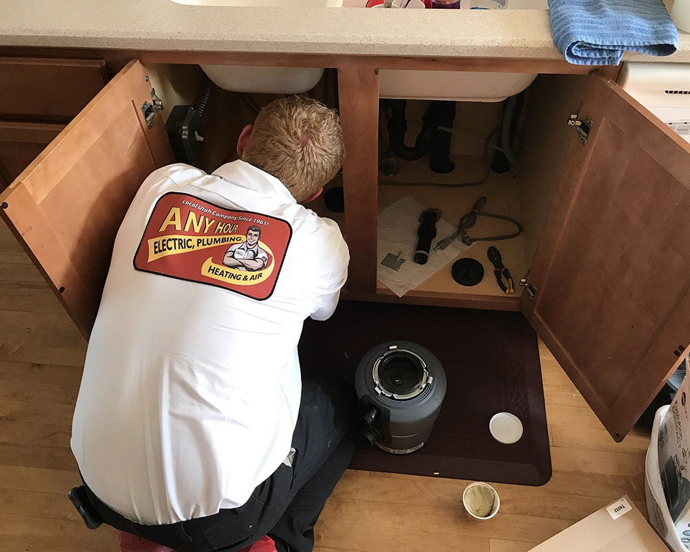 A plumber in an AnyHour Services shirt repairs pipes under a kitchen sink. Tools are scattered around, conveying focus and professionalism.