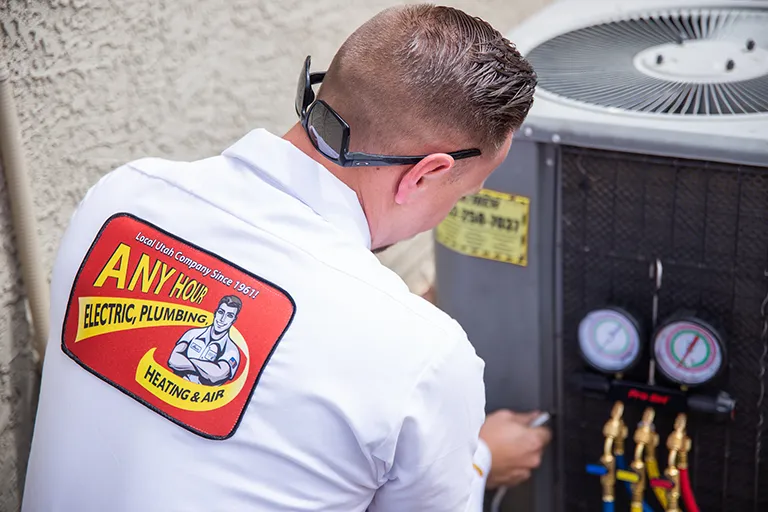 A technician in a white uniform with Any Hour Electric, Plumbing, Heating & Air logo works on an HVAC unit, adjusting gauges with focus and care.