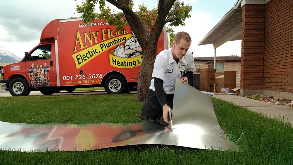 A technician in a white uniform kneels on a lawn, cutting a shiny metal sheet. Nearby, a red service truck for electric, plumbing, and heating is parked.