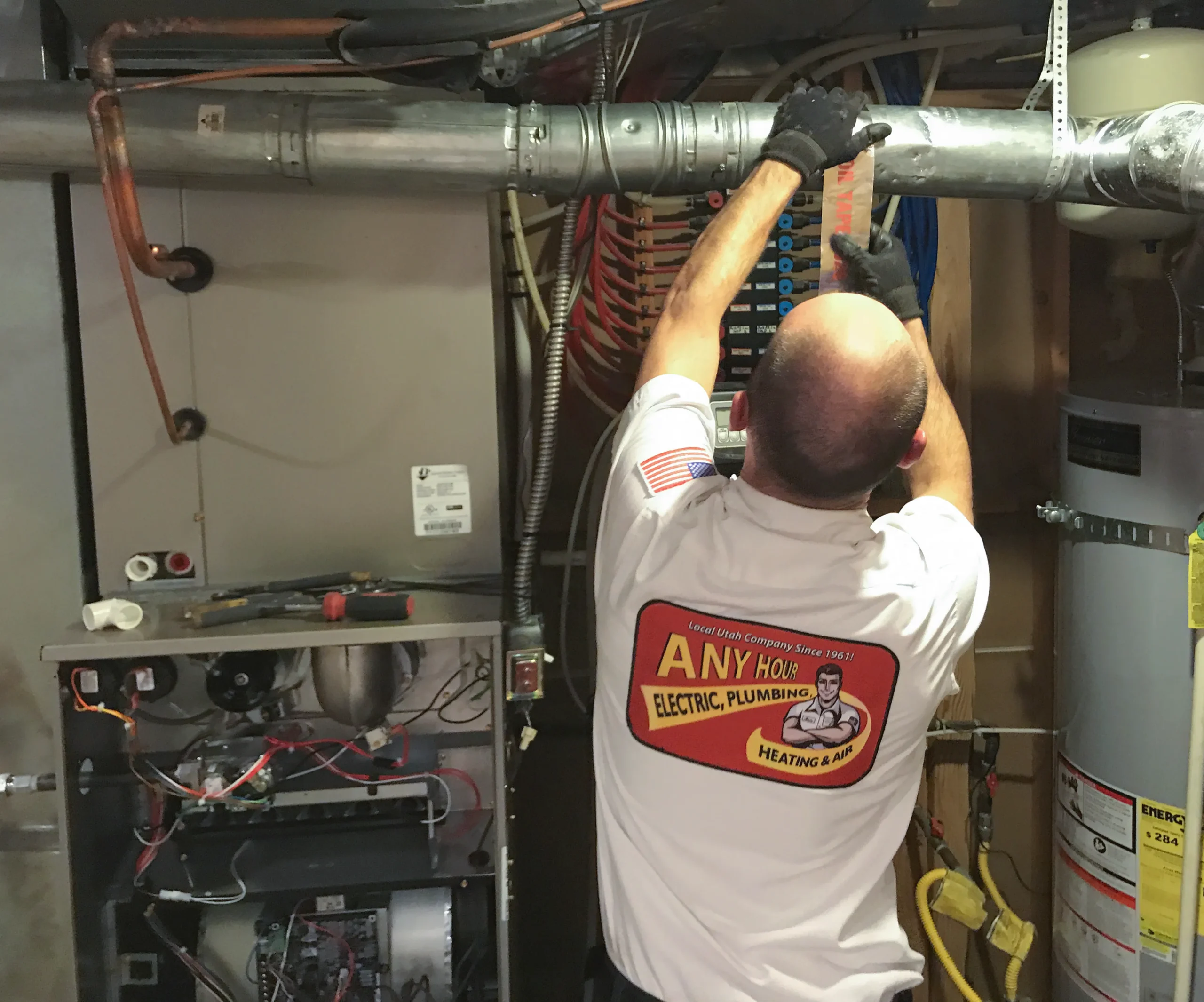 A person in a work uniform labeled Any Hour is inspecting a metal HVAC system in a utility room. The scene conveys professionalism and focus.