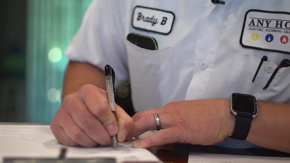 A person in a work uniform is writing on paper at a desk. The shirt has a name tag and company logo. The mood is professional and focused.