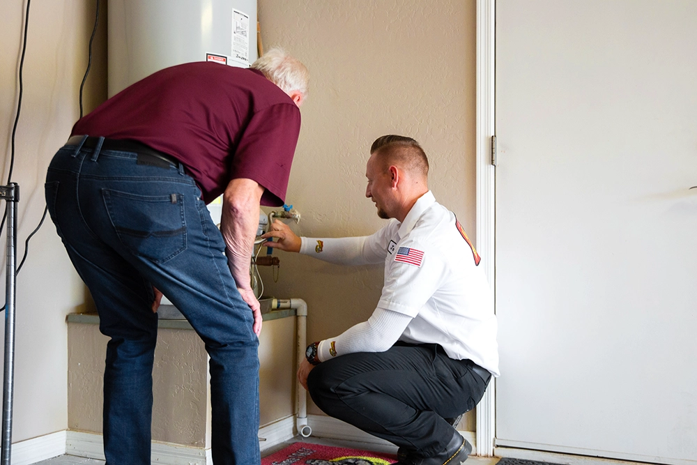 A technician kneels beside a homeowner, explaining a water heater control. The scene conveys professionalism and helpfulness in a home setting.