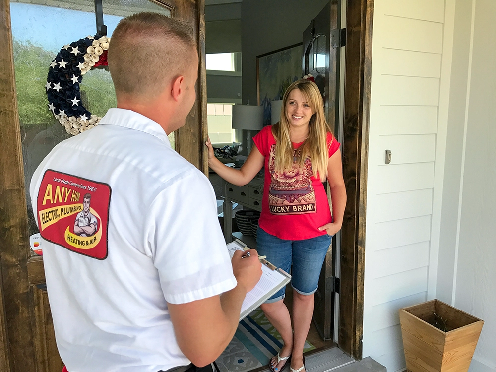 A woman in a red shirt is smiling at a service technician holding a clipboard at her front door. The technician's shirt shows an Electric, Plumbing, Heating & Air logo. The mood is friendly and professional.