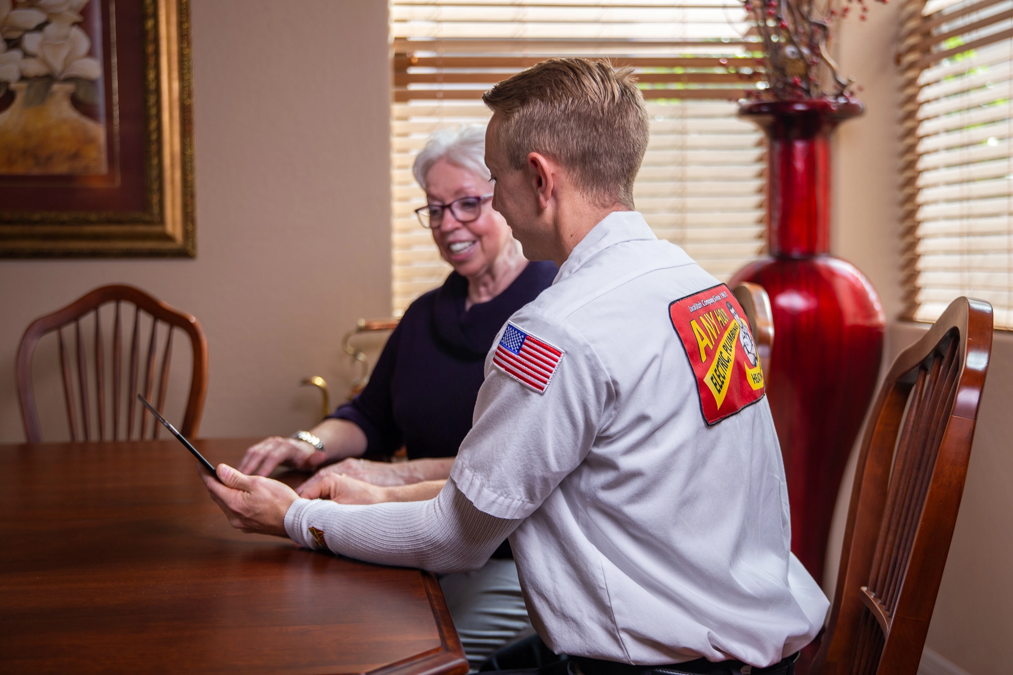 A technician in a uniform sits with a smiling older woman at a wooden table, showing her a tablet. The setting is cozy with warm lighting and decor.