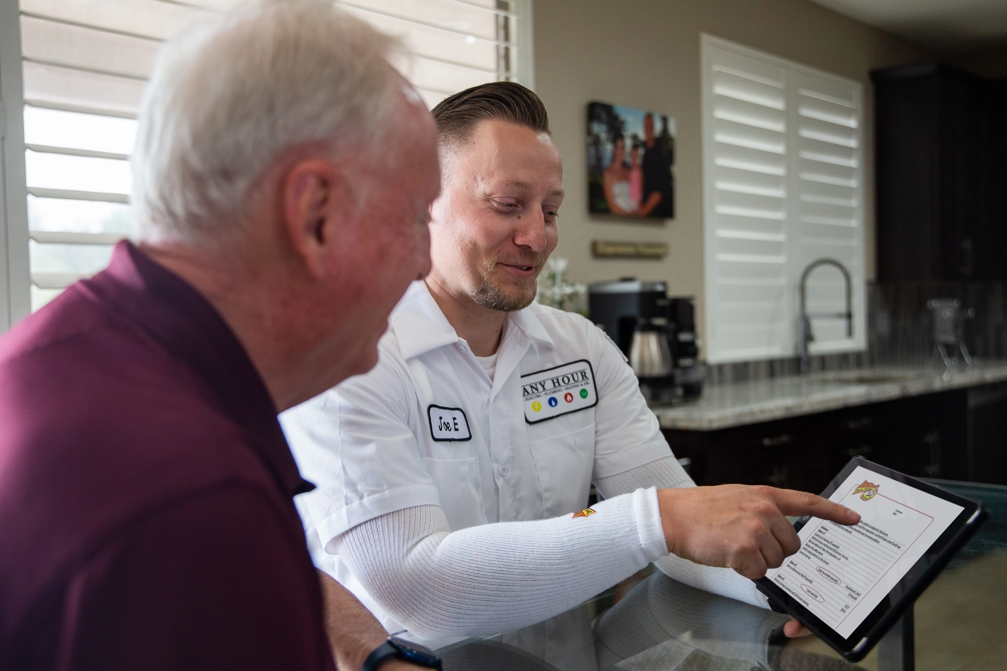 A technician in a white uniform shows an elderly man a document on a tablet in a bright kitchen. Both are engaged and smiling, conveying friendliness.