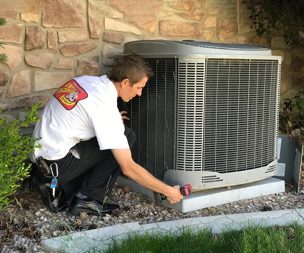 A technician in a uniform crouches outside inspecting an air conditioning unit with a tool. The setting is residential, with a stone wall and foliage.