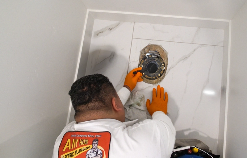 A plumber wearing orange gloves is fixing a toilet flange on a white marble bathroom floor. The mood is focused and professional.