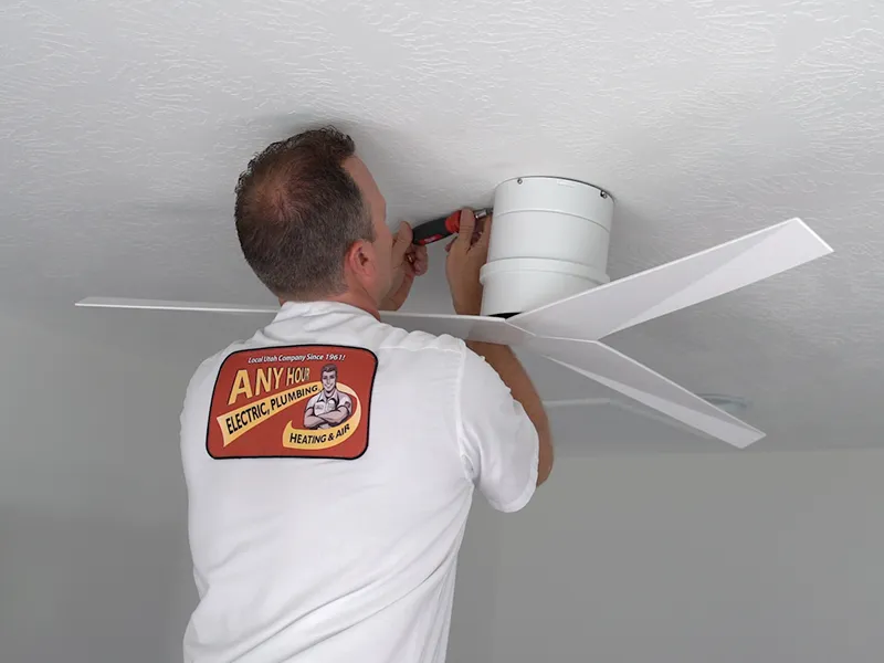 A technician in a branded shirt installs a modern white ceiling fan. He uses tools, focused on completing the task in a brightly lit room.