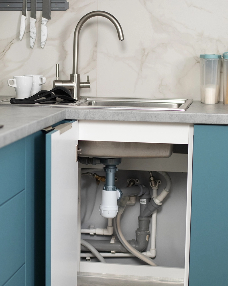 Modern kitchen sink with sleek silver faucet, blue cabinets, and open door revealing plumbing underneath. Counter holds mugs and storage canisters.