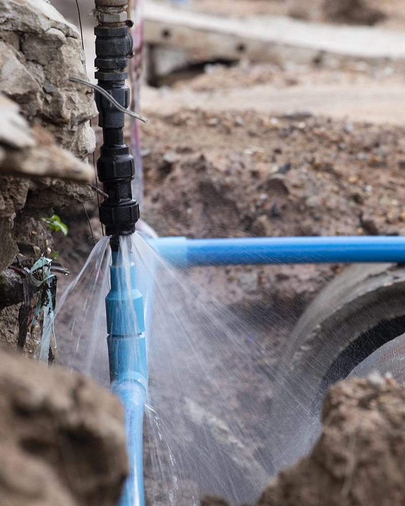 A blue water pipe set in dirt is leaking, spraying water through a crack. The surrounding area appears muddy, highlighting the urgency for repair.