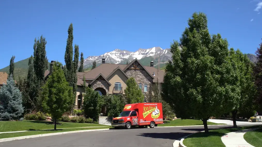 A red AnyHour service truck is parked outside a large stone house with tall trees and a mountain backdrop under a clear blue sky, conveying a serene suburban scene.