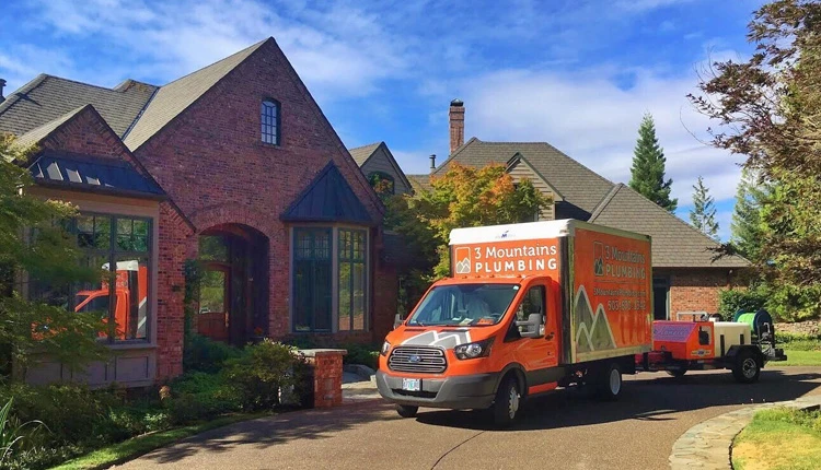 Orange 3 Mountains Plumbing van parked in front of a large red-brick house with a well-maintained garden under a blue sky, conveying reliability.