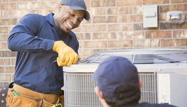 Two technicians work on an outdoor AC unit. One smiles, holding a tool, wearing gloves, a cap, and a blue uniform by a brick wall in a sunny setting.